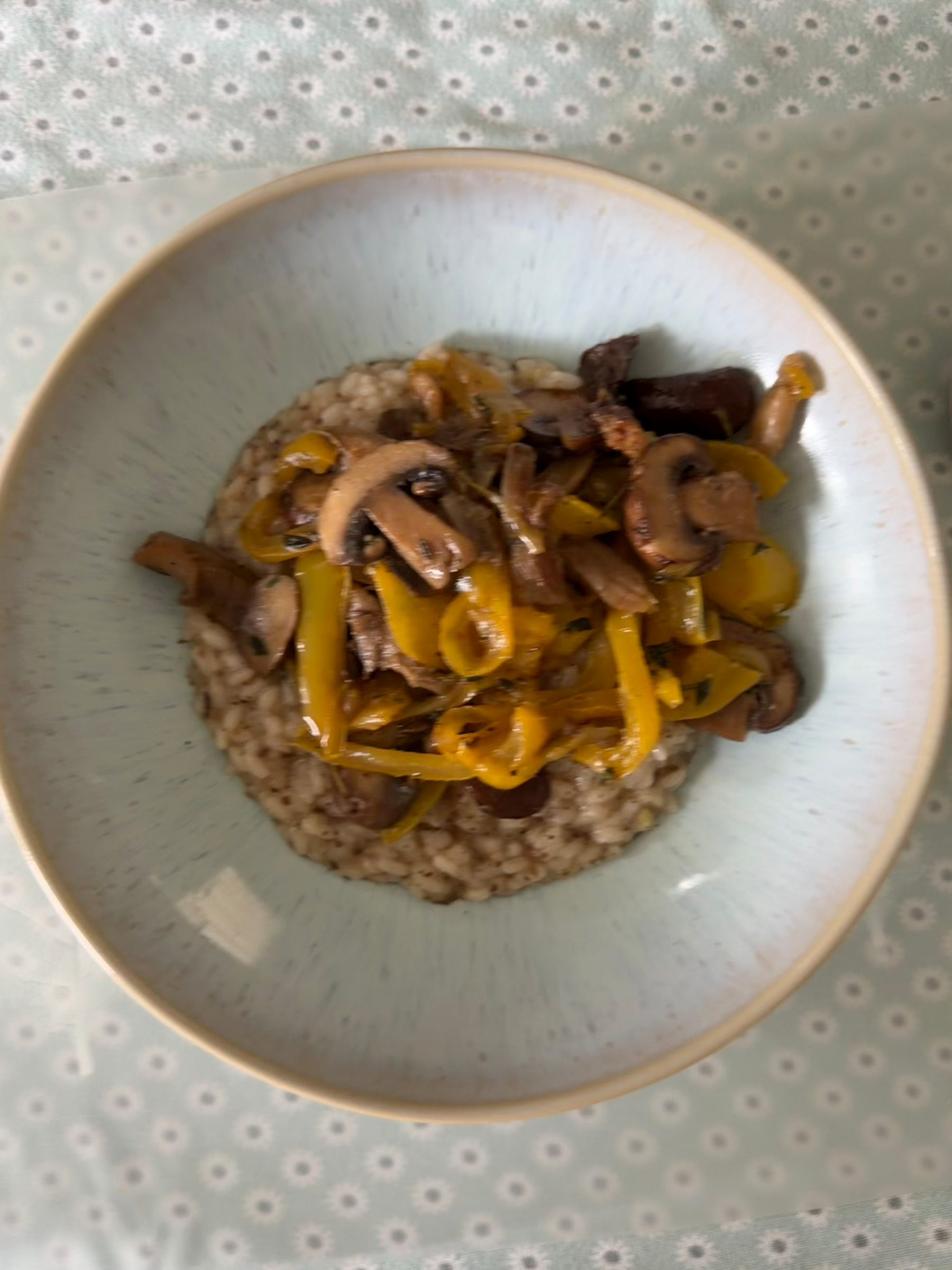 A bowl of risotto topped with sautéed mushrooms and yellow bell peppers is placed on a patterned tablecloth.